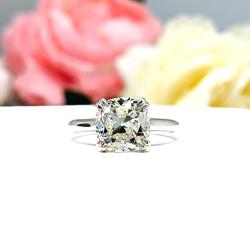 A sterling silver split-prong cathedral engagement ring set with a near colorless old mine cut cubic zirconia. The ring is set against a white background with a blurred image of pink and white flowers in the background.