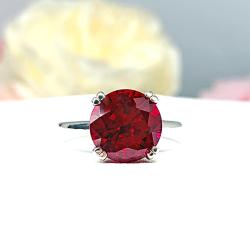 Sterling silver solitaire ring featuring a vibrant round lab-created ruby held in a modern split-prong setting. The rich red gemstone radiates warmth and intensity, its bold color standing out against the cool gleam of polished silver—framed by a soft floral backdrop that enhances its romantic simplicity.