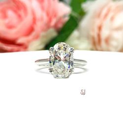 Sterling silver solitaire engagement ring set with a 12x8mm elongated oval near colorless cubic zirconia. The ring features split prongs and a cathedral shank. It is resting on a white surface with soft-focus pink and white flowers in the background.