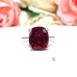 Sterling silver ring featuring a vivid elongated cushion-cut lab created ruby held in a split-prong setting, with soft-focus pink and white flowers in the background and “SJ” initials subtly visible in the corner.