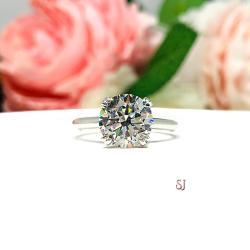Close-up view of a near colorless (G/H/I color) round brilliant cut cubic zirconia solitaire ring in a split prong cathedral setting. The ring is made of sterling silver and features a sleek, minimalist design. The setting is simple yet elegant, allowing the center stone to take center stage. The ring is displayed against a soft, blurred background of pastel-colored flowers, creating a romantic and delicate ambiance.