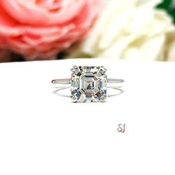 Sterling silver 8mm near colorless (G/H color) asscher cut cubic zirconia solitaire engagement ring. The ring is resting on a white surface with a background of pink and white flowers.