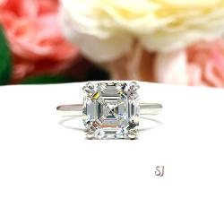 Sterling silver 9mm near colorless (G/H color) asscher cut cubic zirconia solitaire engagement ring. The ring is resting on a white surface with a background of pink and white flowers.