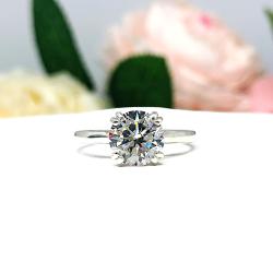 Close-up view of a near colorless (G/H color) round brilliant cut cubic zirconia solitaire ring in a split prong cathedral setting. The ring is made of sterling silver and features a sleek, minimalist design. The setting is simple yet elegant, allowing the center stone to take center stage. The ring is displayed against a soft, blurred background of pastel-colored flowers, creating a romantic and delicate ambiance.