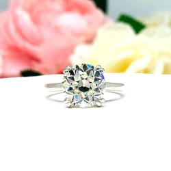 A romantic close-up of a 10mm (4 ct) old European cut near-colorless CZ solitaire engagement ring. Set in a sterling silver split prong cathedral-style mount, the ring gleams with vintage-inspired elegance. Soft pink and white flowers blur in the background, adding a dreamy, bridal ambiance.