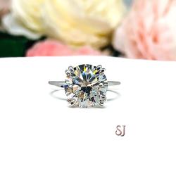 Close-up view of a 10mm near colorless (G/H/I color) round brilliant cut cubic zirconia solitaire ring in a split prong cathedral setting. The ring is made of sterling silver and features a sleek, minimalist design. The setting is simple yet elegant, allowing the center stone to take center stage. The ring is displayed against a soft, blurred background of pastel-colored flowers, creating a romantic and delicate ambiance.
